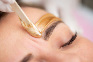 Applying Gold Coloured Wax with Spatula on Woman's Eyebrow