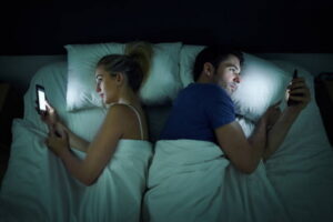 Shot of a young couple using their mobile phones in bed at night back to back
