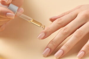 A close-up photograph of a hand with a subtle light pink manicure receiving a single, clear drop of golden-coloured cuticle oil from a small glass dropper bottle