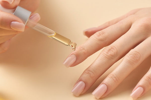 A close-up photograph of a hand with a subtle light pink manicure receiving a single, clear drop of golden-coloured cuticle oil from a small glass dropper bottle