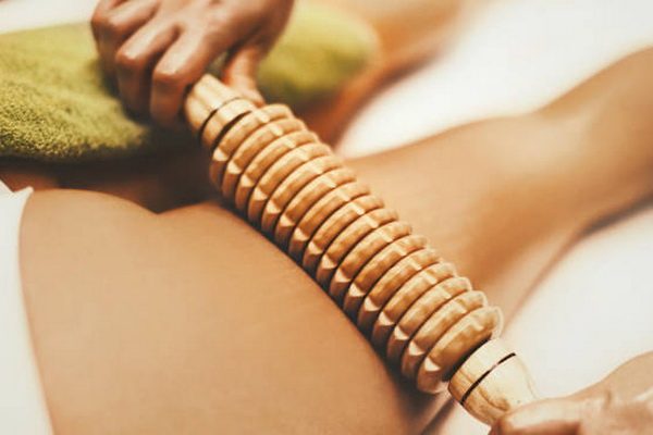 Close-up of a massage therapist using a wooden roller tool on a women's leg to stimulate and improve blood circulation.